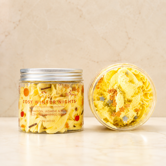 Two clear jars of Cosy Winter Nights scented whipped soap on a light marble surface against a beige marble wall. One jar is closed with a silver lid, showing the label, and the other is open to show the pale yellow whipped soap inside with colorful star flecks.