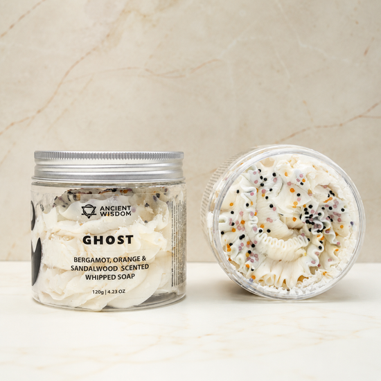 A jar of Ghost Bergamot, Orange & Sandalwood Scented Whipped Soap shown next to its lid, which is open to reveal the whipped soap inside. The soap is white with decorative black, orange, and yellow sprinkles, set against a light beige marble background.