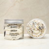 A jar of Ghost Bergamot, Orange & Sandalwood Scented Whipped Soap shown next to its lid, which is open to reveal the whipped soap inside. The soap is white with decorative black, orange, and yellow sprinkles, set against a light beige marble background.