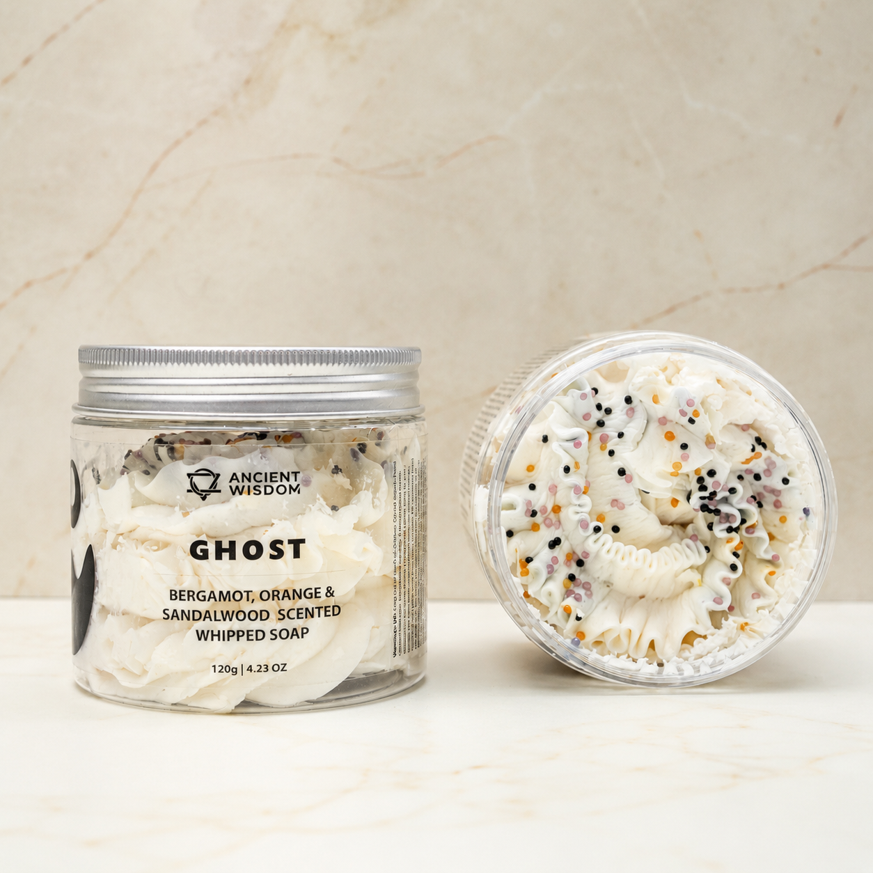 A jar of Ghost Bergamot, Orange & Sandalwood Scented Whipped Soap shown next to its lid, which is open to reveal the whipped soap inside. The soap is white with decorative black, orange, and yellow sprinkles, set against a light beige marble background.