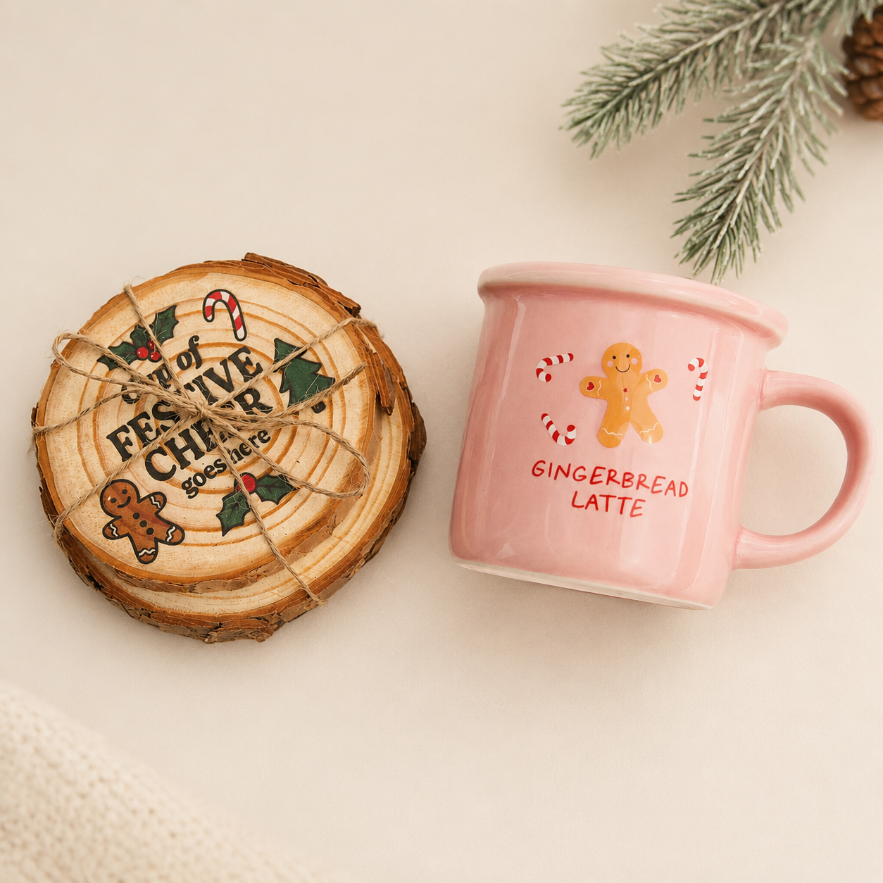 A pink ceramic mug with a gingerbread man illustration and the words 'GINGERBREAD LATTE' sits next to a round wooden coaster printed with 'FESTIVE CHEER' and holiday designs, styled on a light beige surface with pine branches in the corner. Gingerbread Latte Mug & Coaster Set