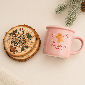 A pink ceramic mug with a gingerbread man illustration and the words 'GINGERBREAD LATTE' sits next to a round wooden coaster printed with 'FESTIVE CHEER' and holiday designs, styled on a light beige surface with pine branches in the corner. Gingerbread Latte Mug & Coaster Set