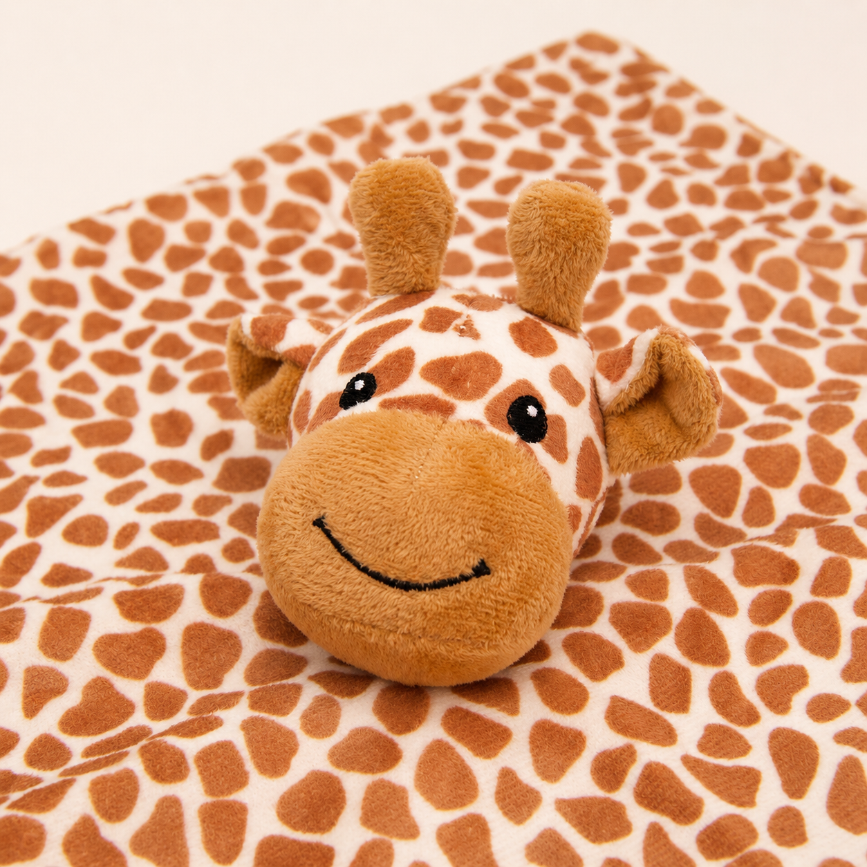 A plush giraffe toy resting on a soft blanket with a matching giraffe print pattern, photographed against a plain light background.