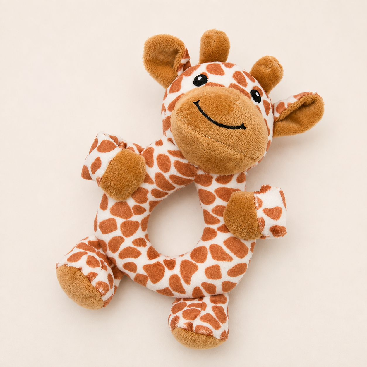 A plush giraffe baby teething ring toy with a soft white fabric body, warm brown spots, and a smiling embroidered face, shown on a plain light beige background.