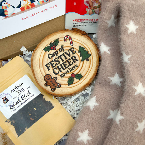 A styled gift box contents featuring a round wooden slice coaster with a festive Christmas design and the text 'Cup of FESTIVE CHEER 2025 here', alongside a bag of 'Artisan Tea Refresh Black', a box of 'Happy New Year' tea, and a soft fuzzy blanket with white stars.