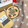A styled gift box contents featuring a round wooden slice coaster with a festive Christmas design and the text 'Cup of FESTIVE CHEER 2025 here', alongside a bag of 'Artisan Tea Refresh Black', a box of 'Happy New Year' tea, and a soft fuzzy blanket with white stars.