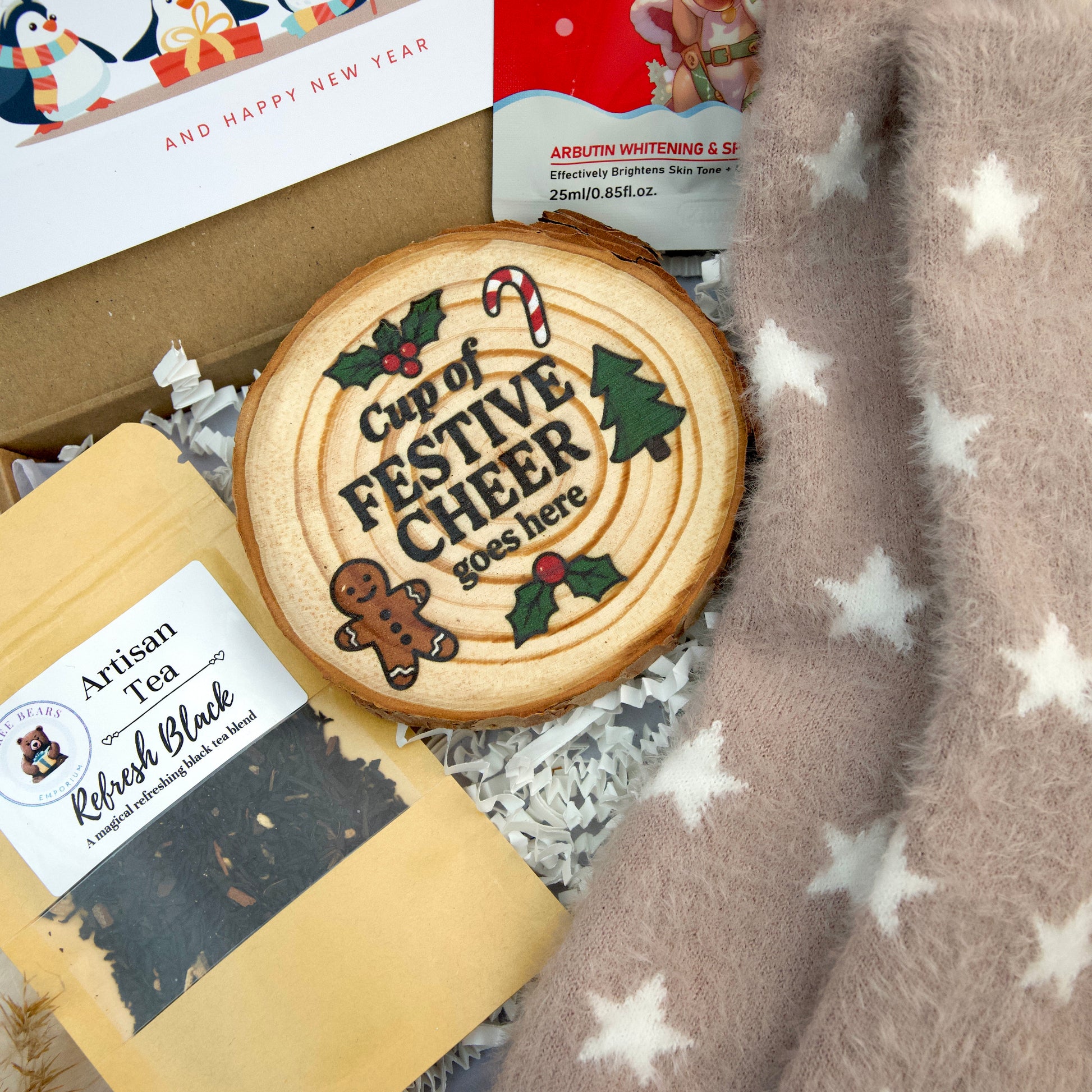 A styled gift box contents featuring a round wooden slice coaster with a festive Christmas design and the text 'Cup of FESTIVE CHEER 2025 here', alongside a bag of 'Artisan Tea Refresh Black', a box of 'Happy New Year' tea, and a soft fuzzy blanket with white stars.